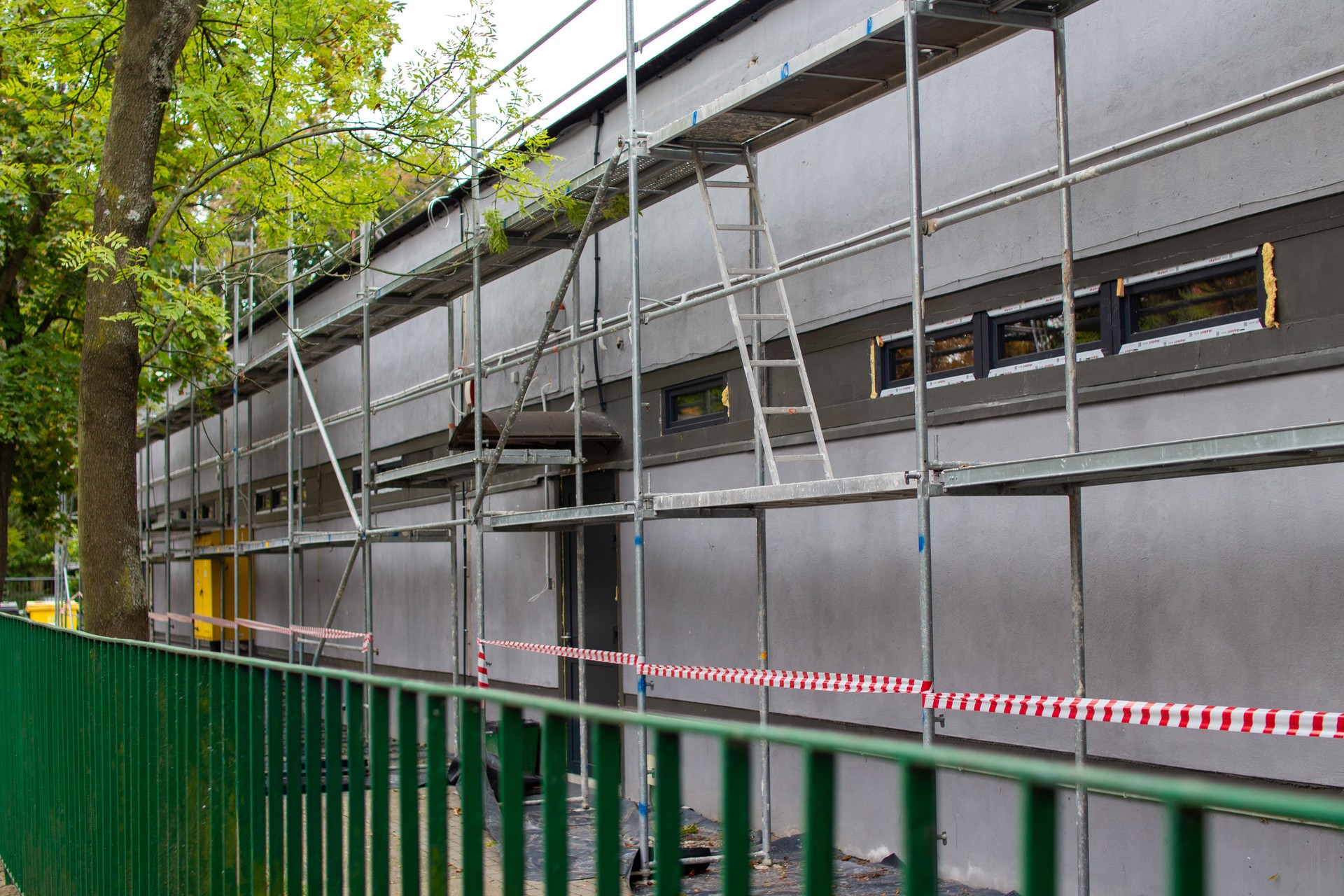 Construction work takes place on a building under renovation with scaffolding and fencing in a green urban setting