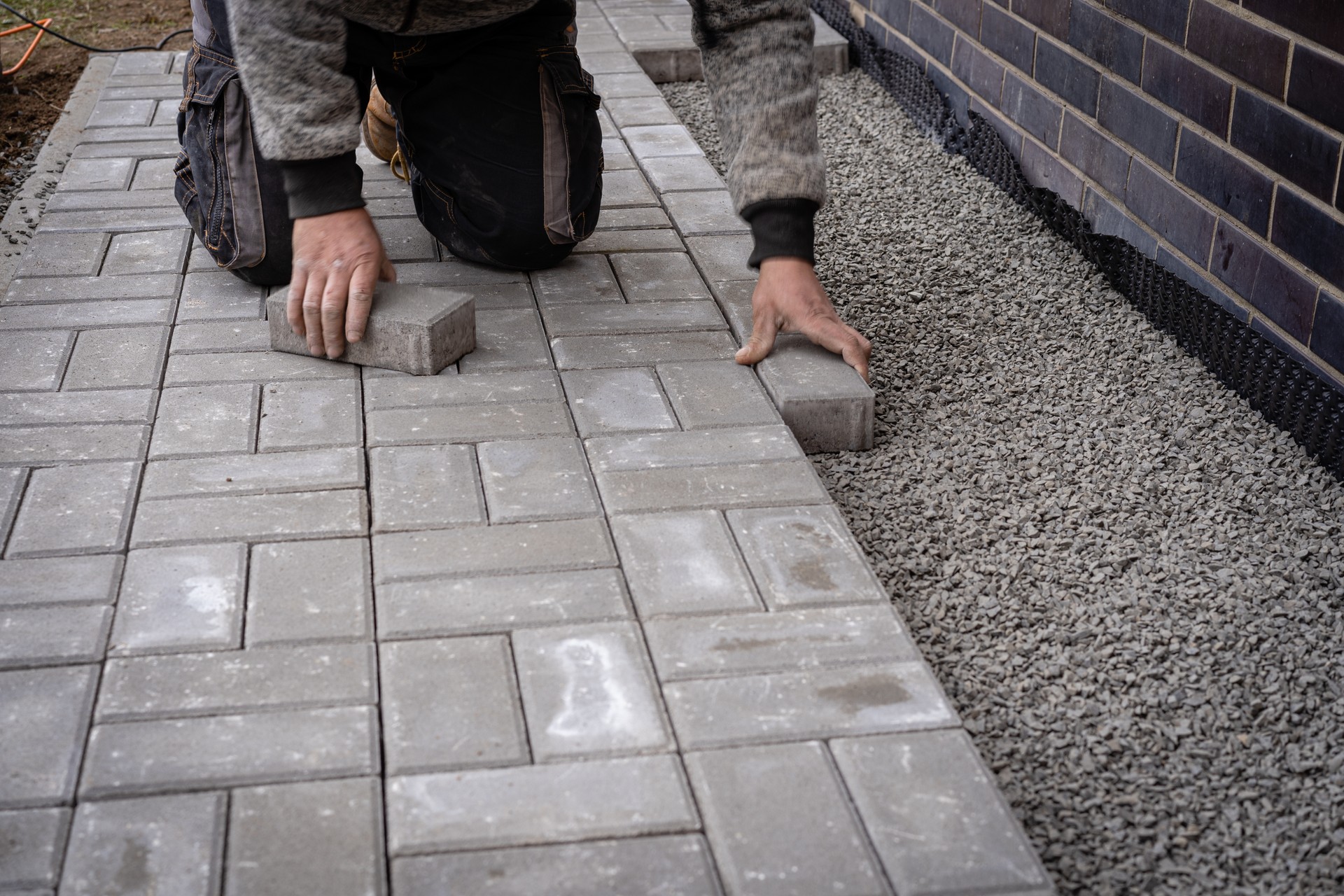 Unidentified worker is laying gray concrete paving slabs in house courtyard. Professional bricklayer is installing new tiles or slabs for walkway, sidewalk or patio on leveled gravel foundation base